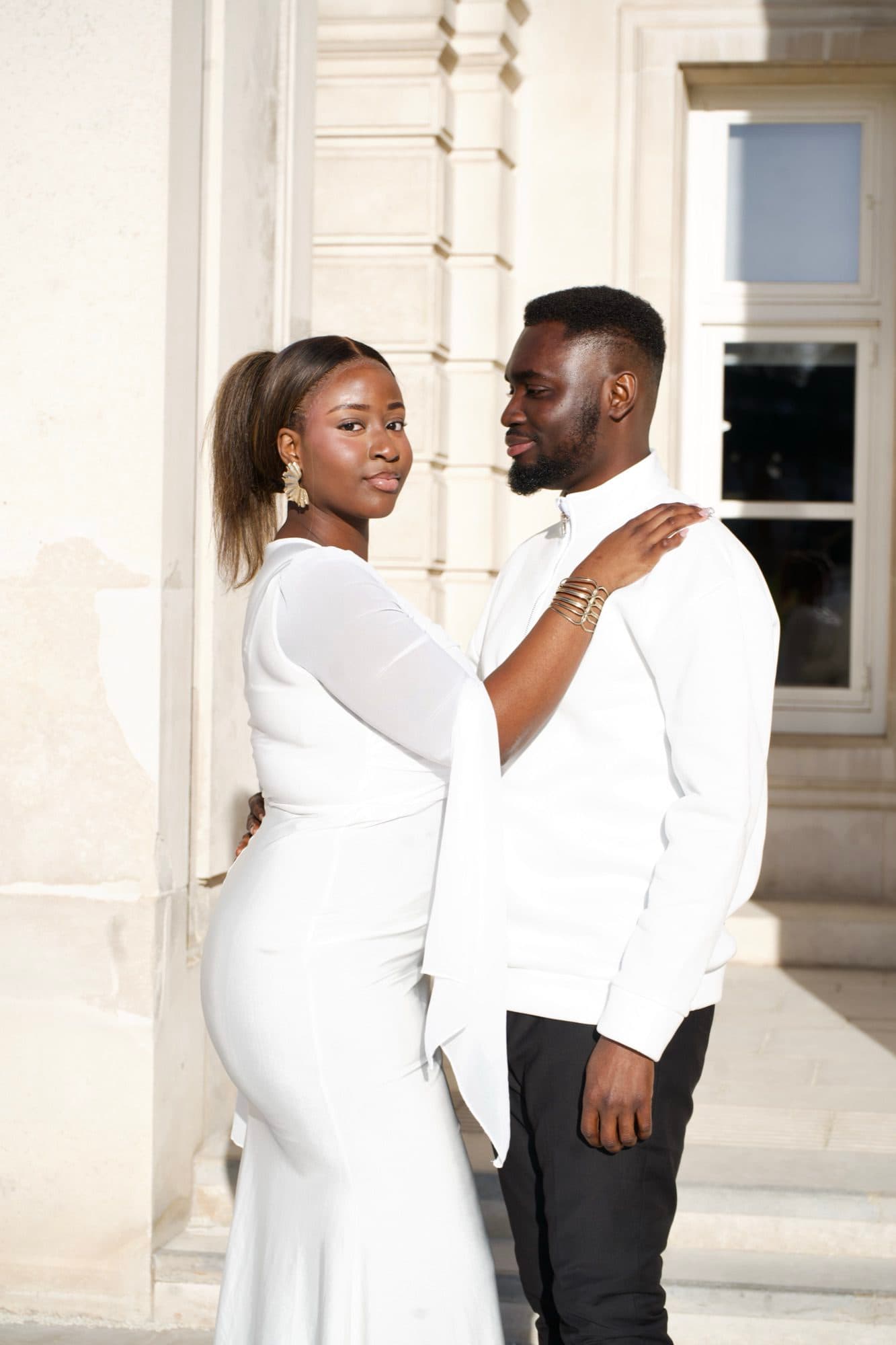 A couple dressed in white, the woman with a hand on the man's shoulder, photographed against tall stone columns