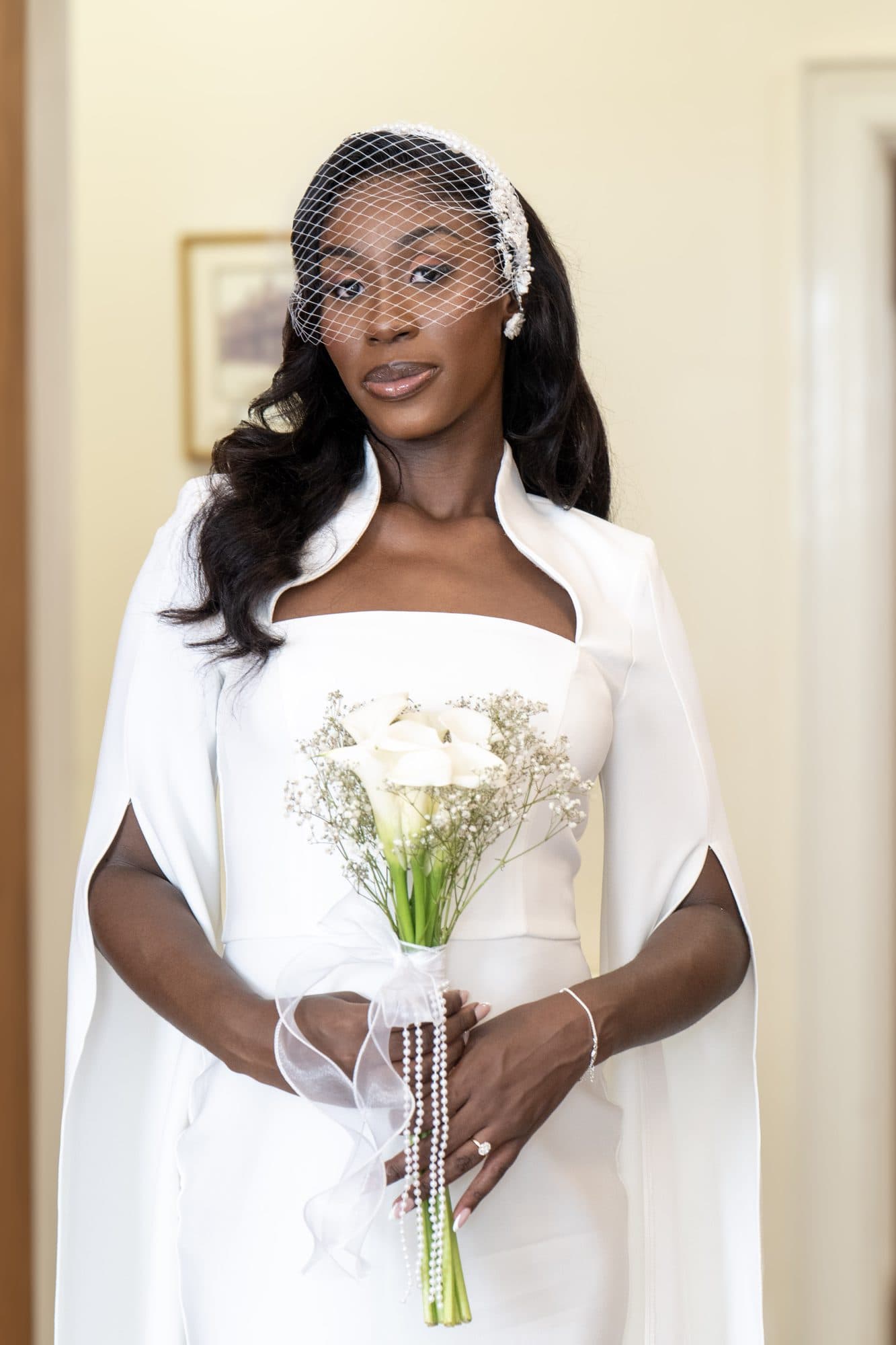 A woman in a white dress with cape sleeves and a birdcage veil, holding a bouquet of white calla lilies