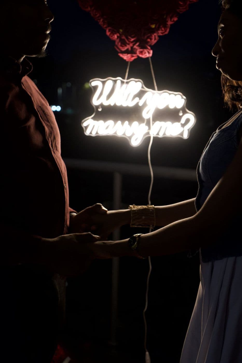 A neon sign reading “will you marry me” glowing behind two silhouetted figures.