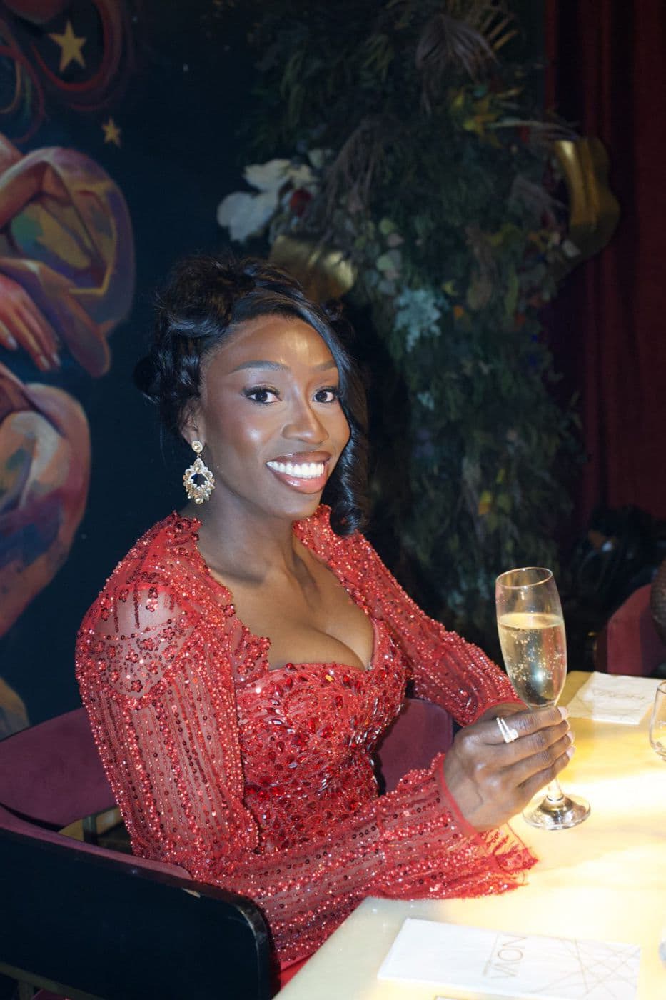 A woman in red seated at a table with a champagne glass, a floral backdrop behind her.