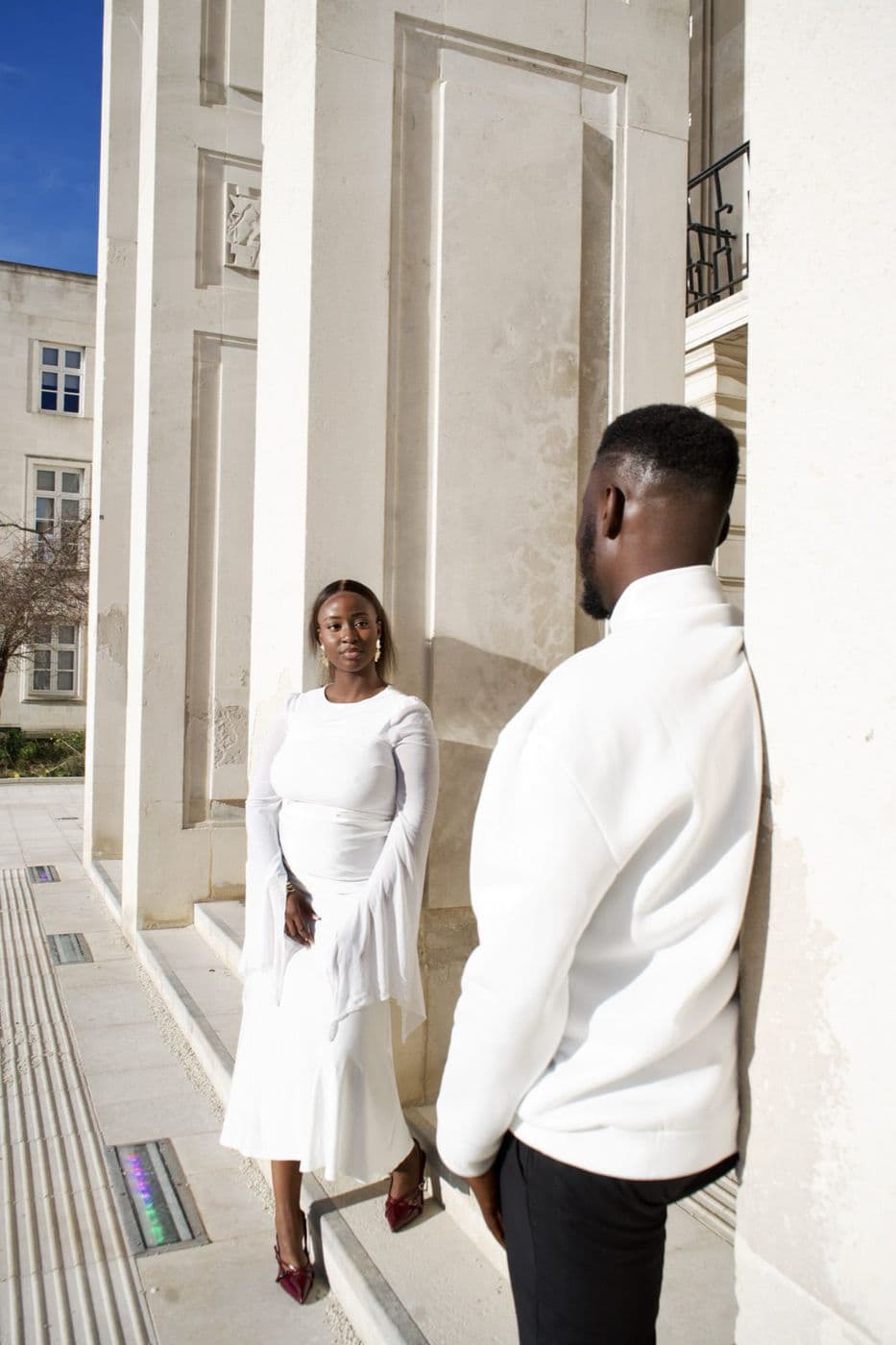 A couple facing each other between stone columns, one in a long white dress.