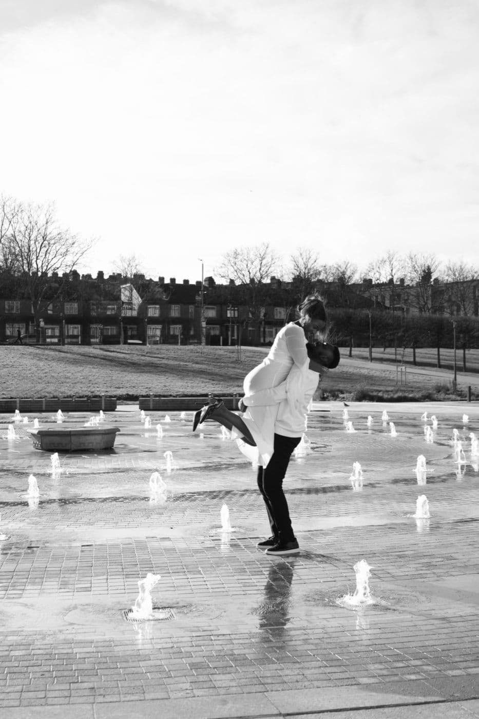 Black and white. A person mid-step among low fountains on a wet plaza.