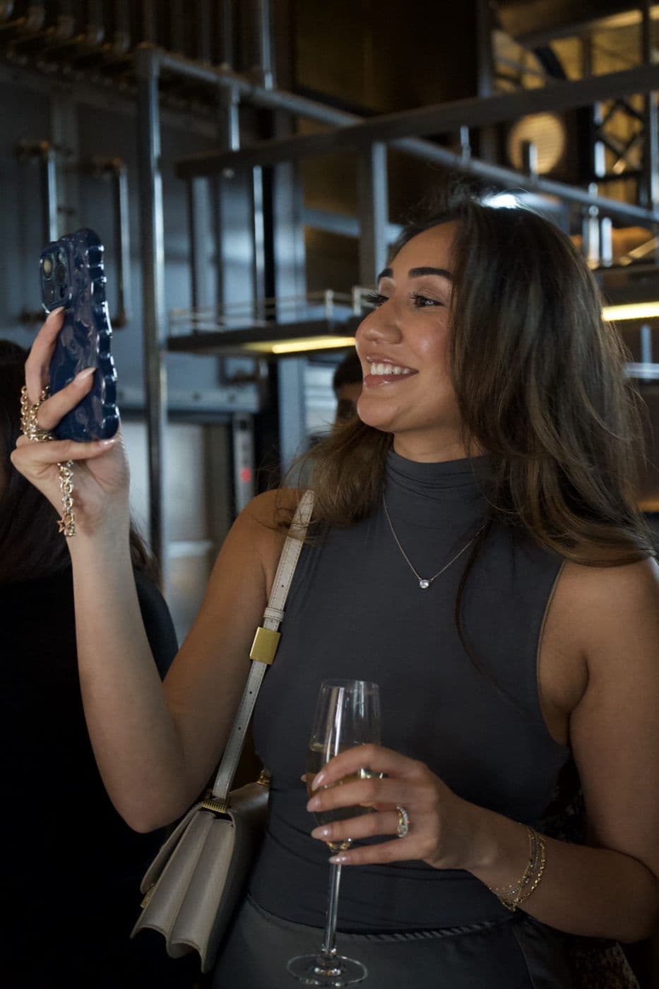 A woman holding a phone and a champagne flute, smiling, indoors.