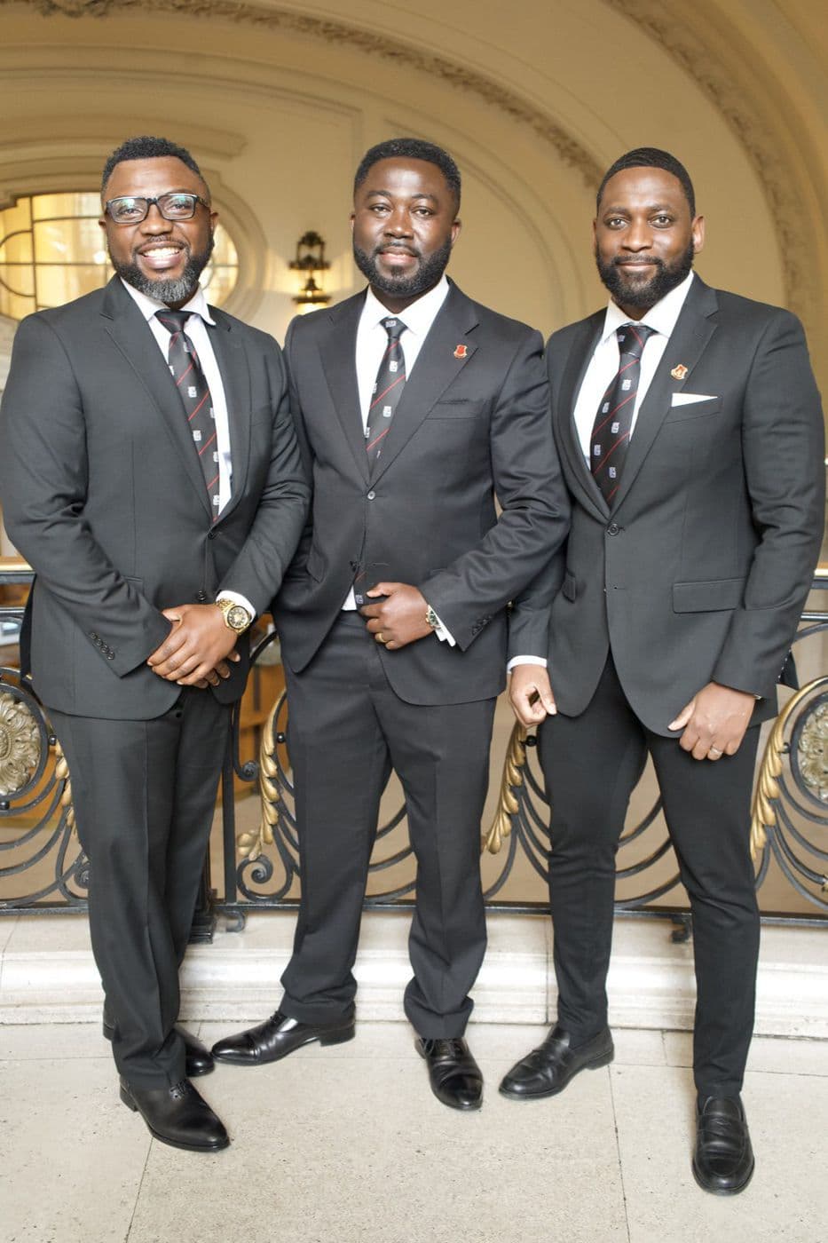 Three men in dark suits standing together beneath a stone arch.