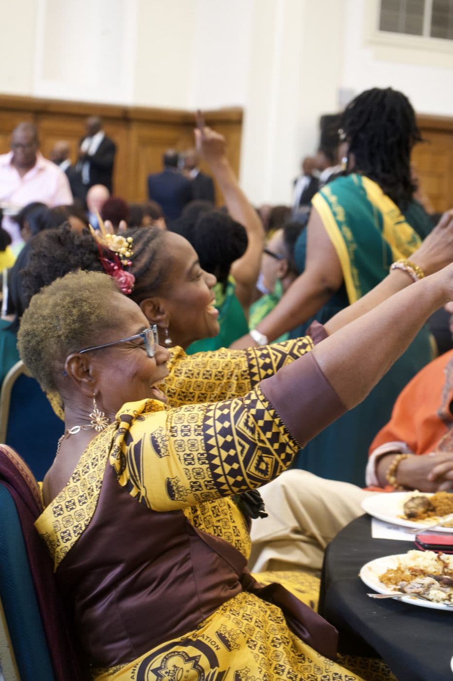 An older woman in patterned yellow and purple raising her arm at a seated gathering.