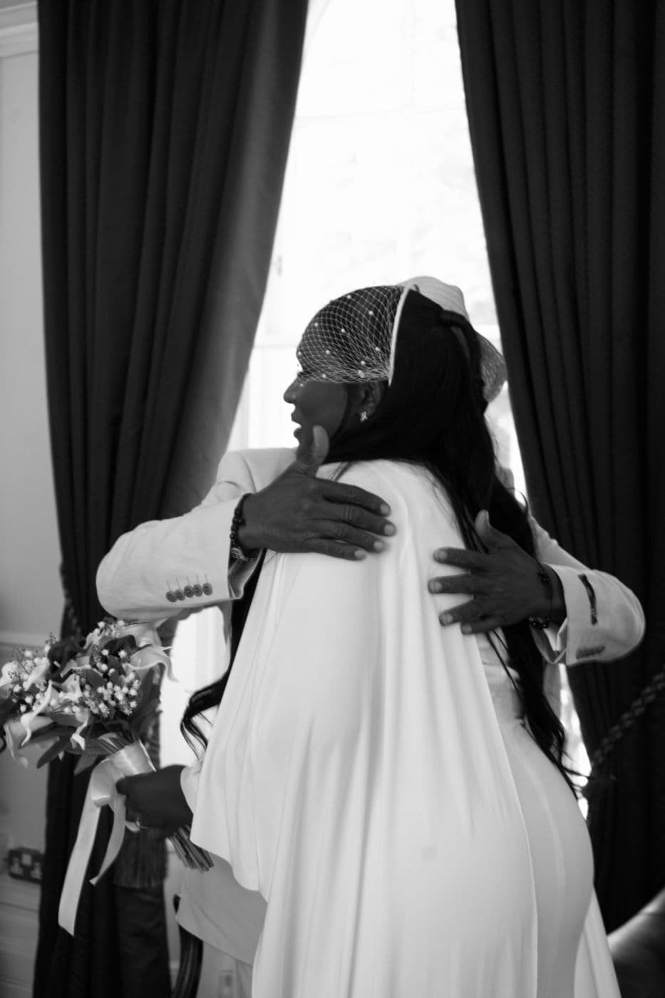 Black and white. Two people embracing, one in a long veil holding a bouquet.