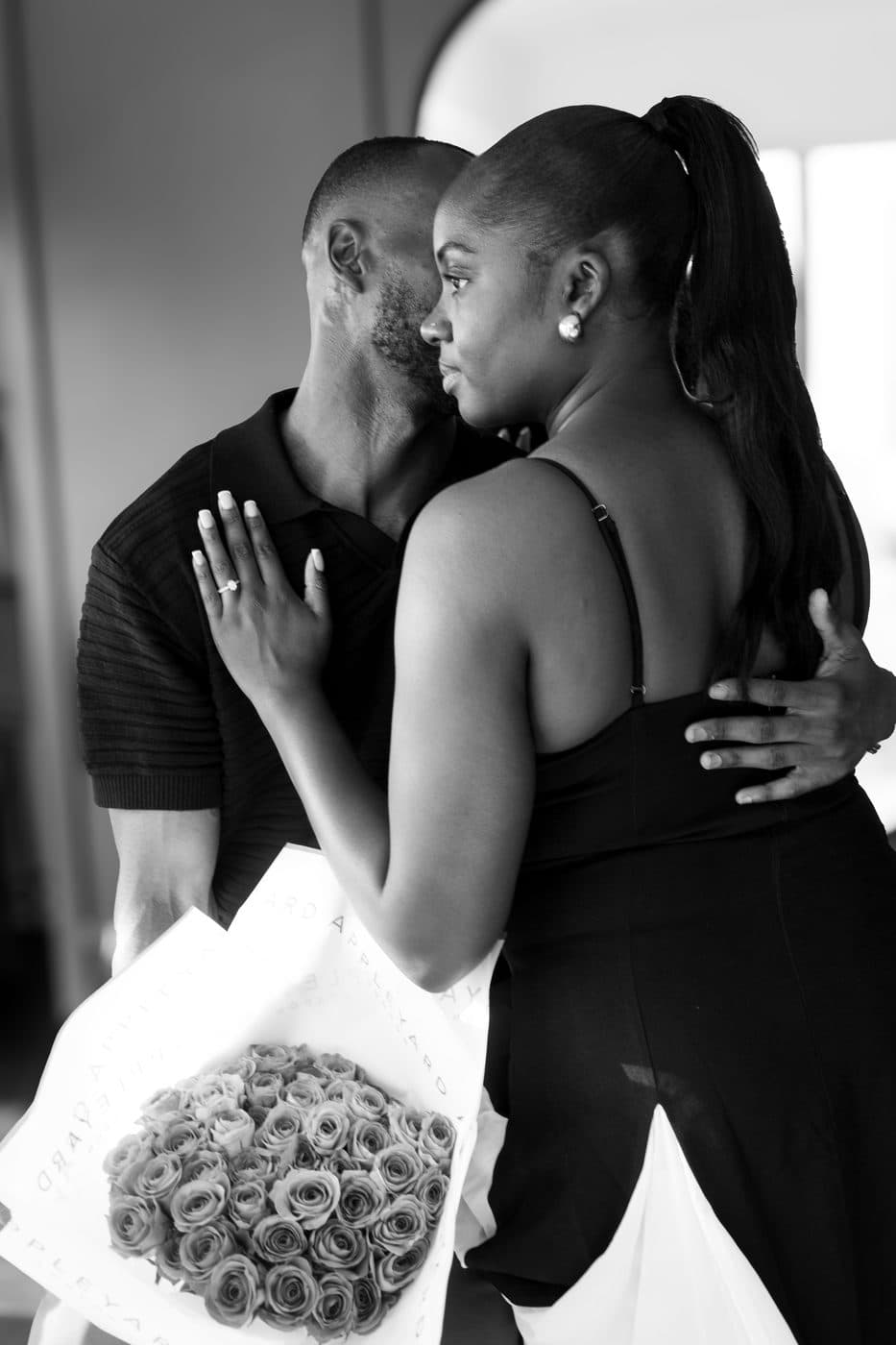 Black and white. A couple embracing closely, a ringed hand resting on the man's chest, a bouquet of roses at her side.