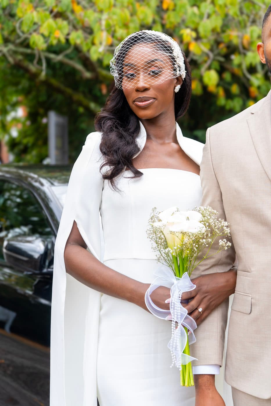 A woman in a white dress with cape sleeves and a birdcage veil, holding a bouquet of white flowers, beside a man in a beige suit.
