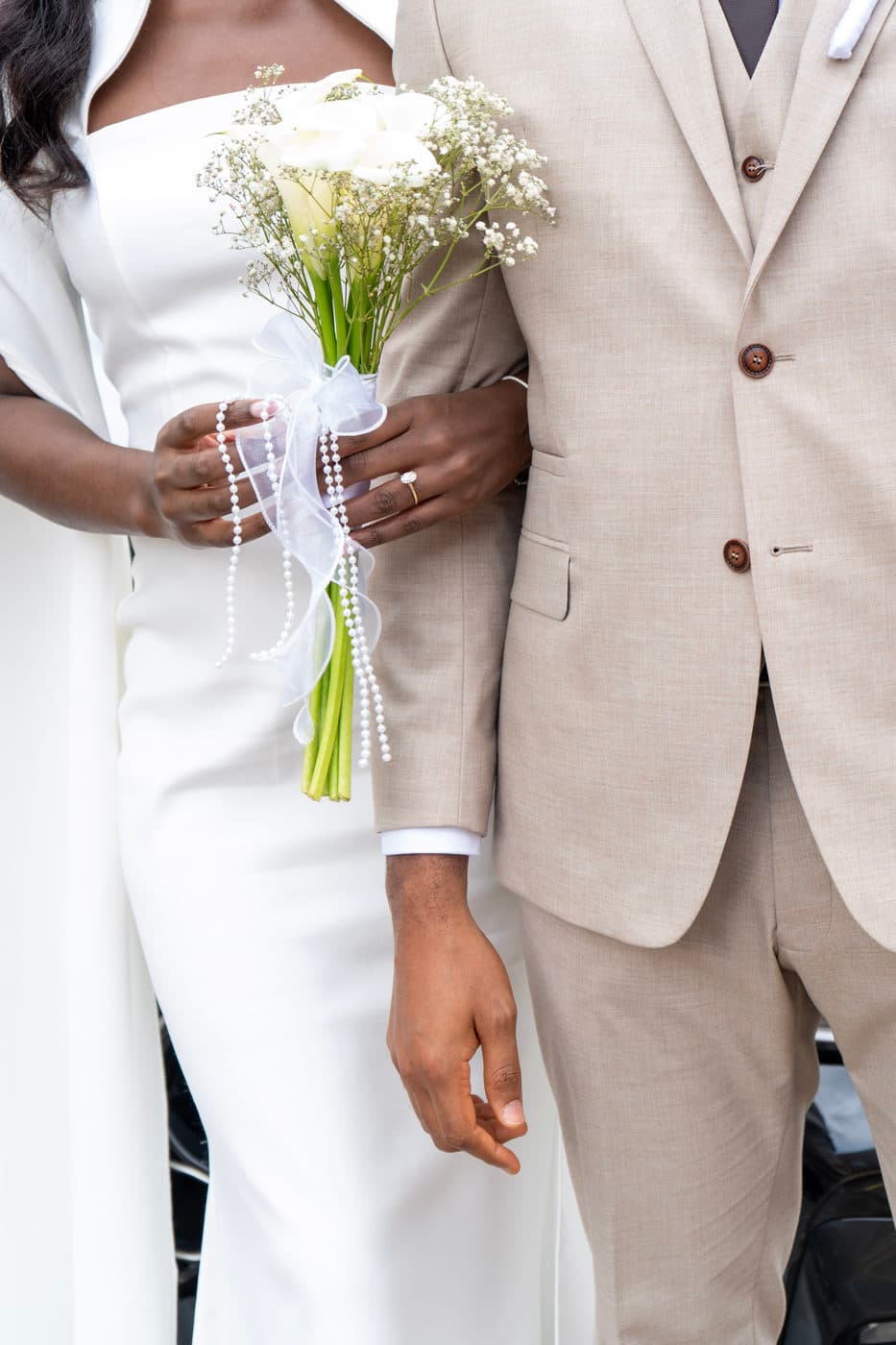 Close on hands: a woman in white holding a bouquet of flowers tied with pearls, resting against a man's beige suit sleeve.