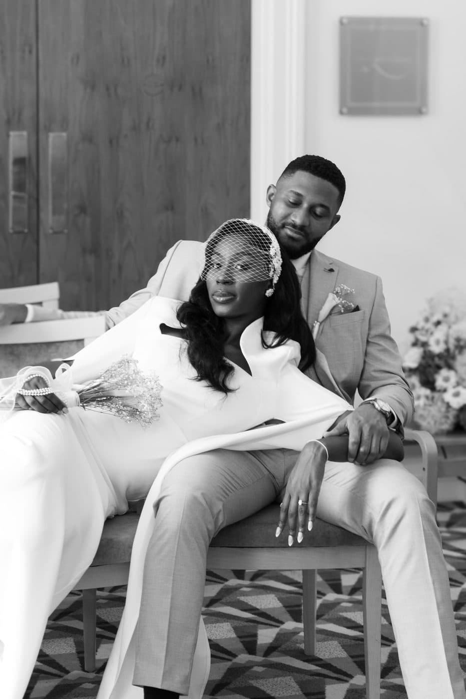 Black and white. A woman in a white dress reclining across a man's lap on a chair, both looking toward camera.