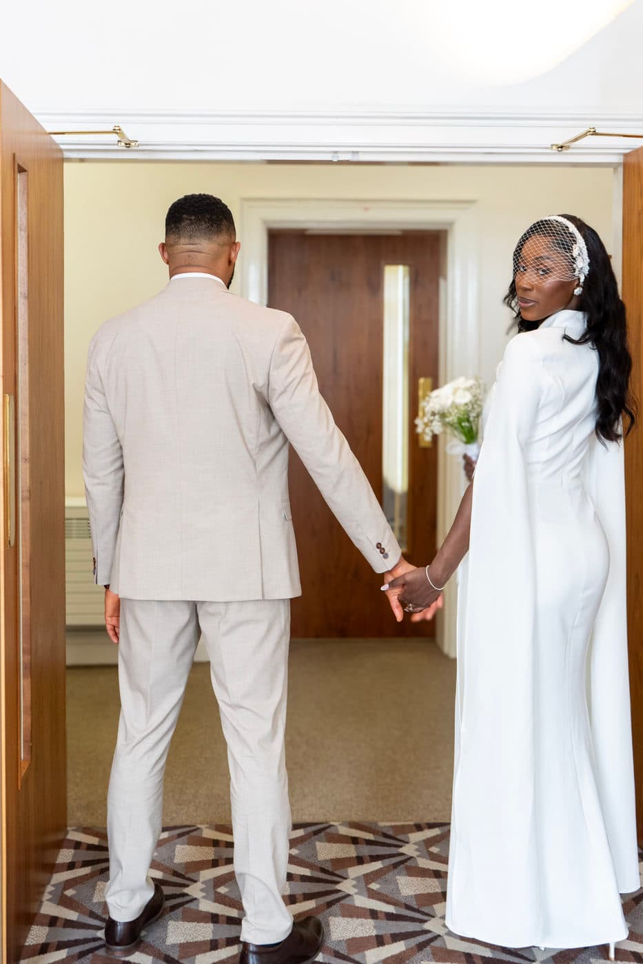 A woman in a white caped dress looking back over her shoulder while walking hand in hand with a man in a beige suit through a doorway.