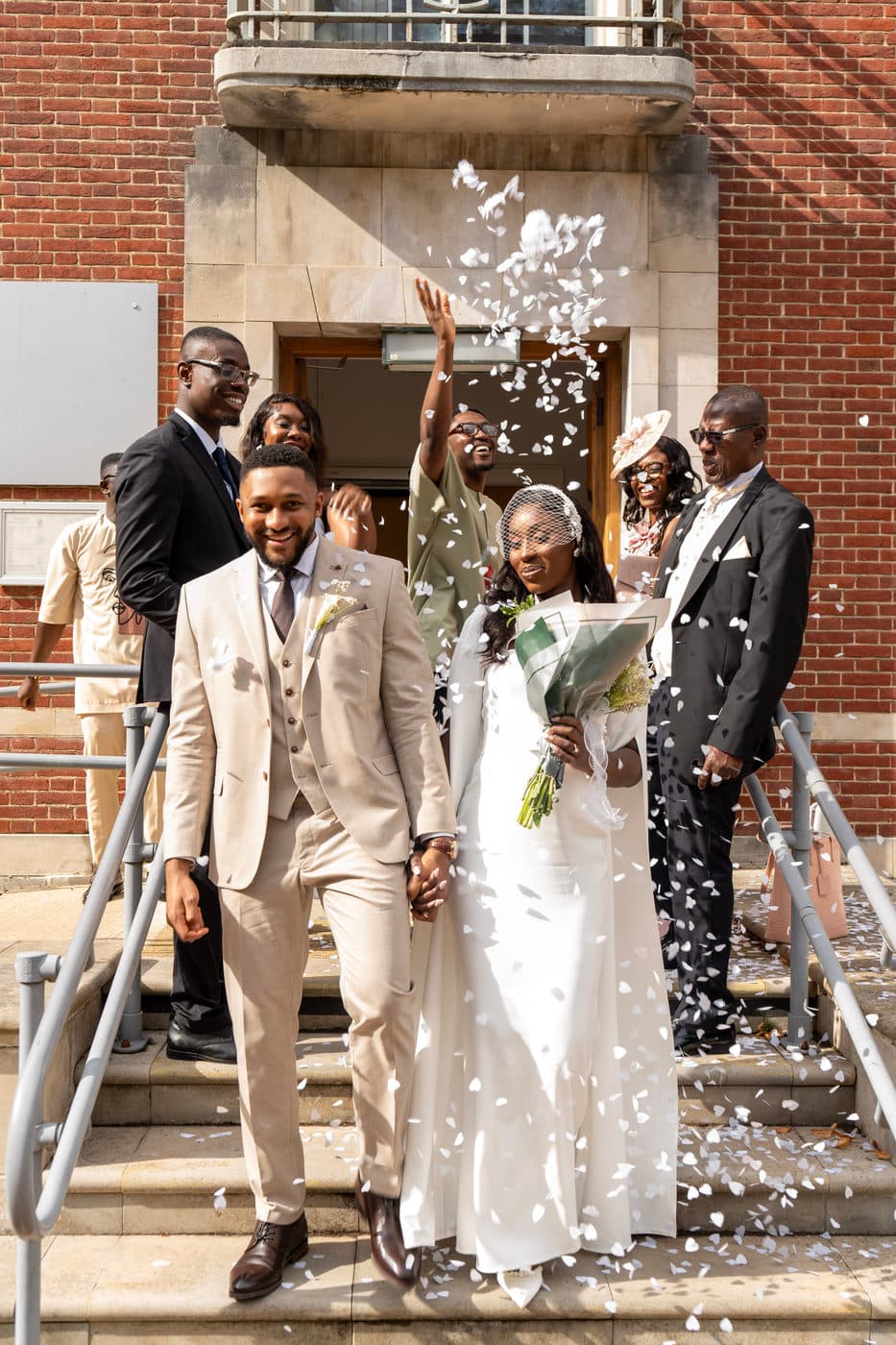 A couple walking down stone steps through falling confetti, surrounded by smiling guests.