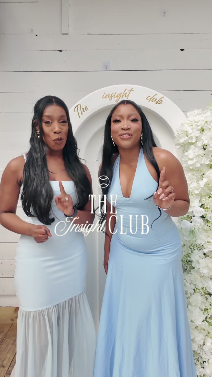 Two women in pale blue dresses at a branded panel event, in front of a floral arch